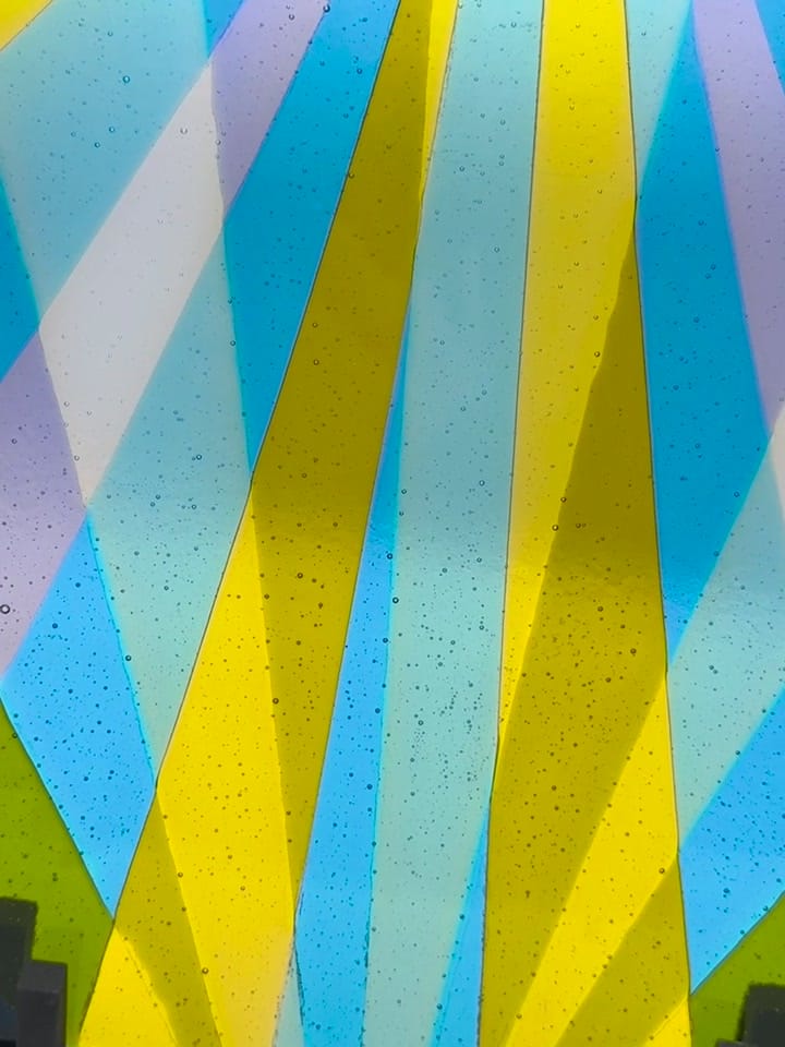Fused-glass weaving panel with diagonal rays of blue, yellow and lavender, backlit so the color glows through